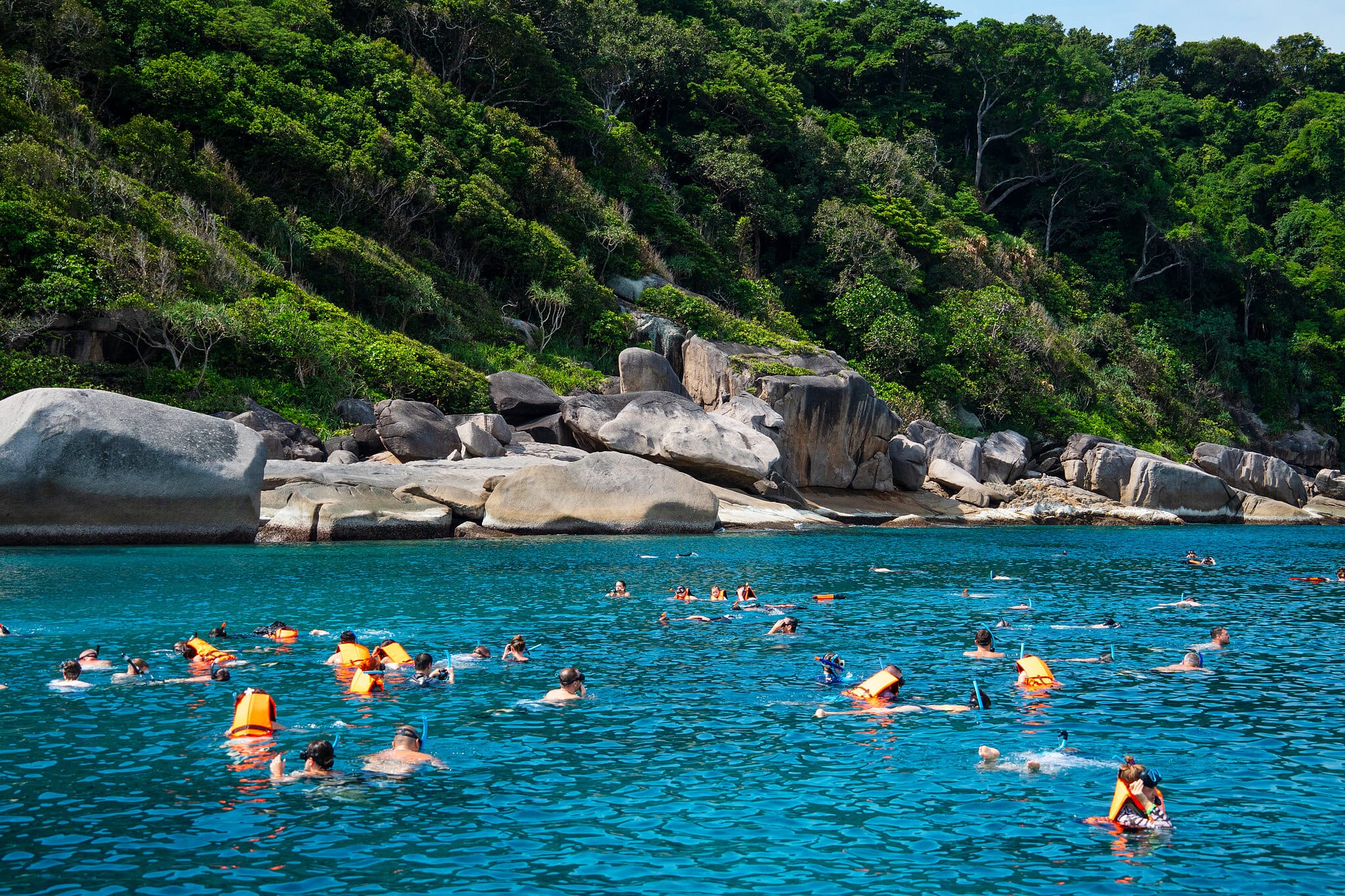 Snorkelers in orange life vests swimming near granite boulders in the clear sea of the Similan Islands