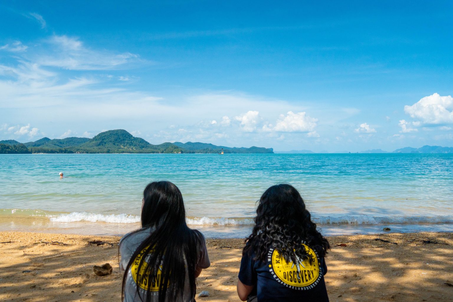 Two Khao Lak Land Discovery team members in company T-shirts sitting on a beach, looking out to sea