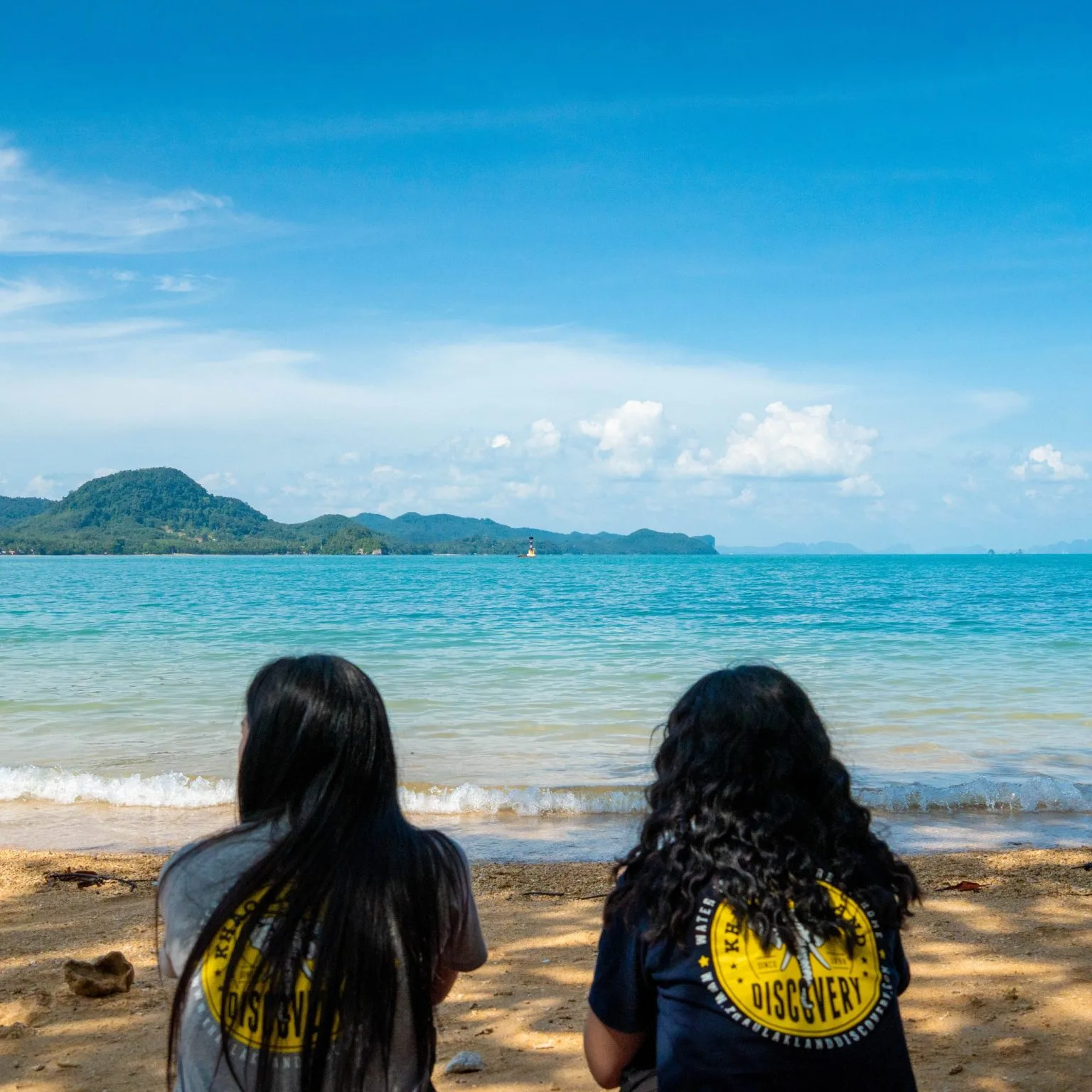 Two Khao Lak Land Discovery team members in company T-shirts sitting on a beach, looking out to sea