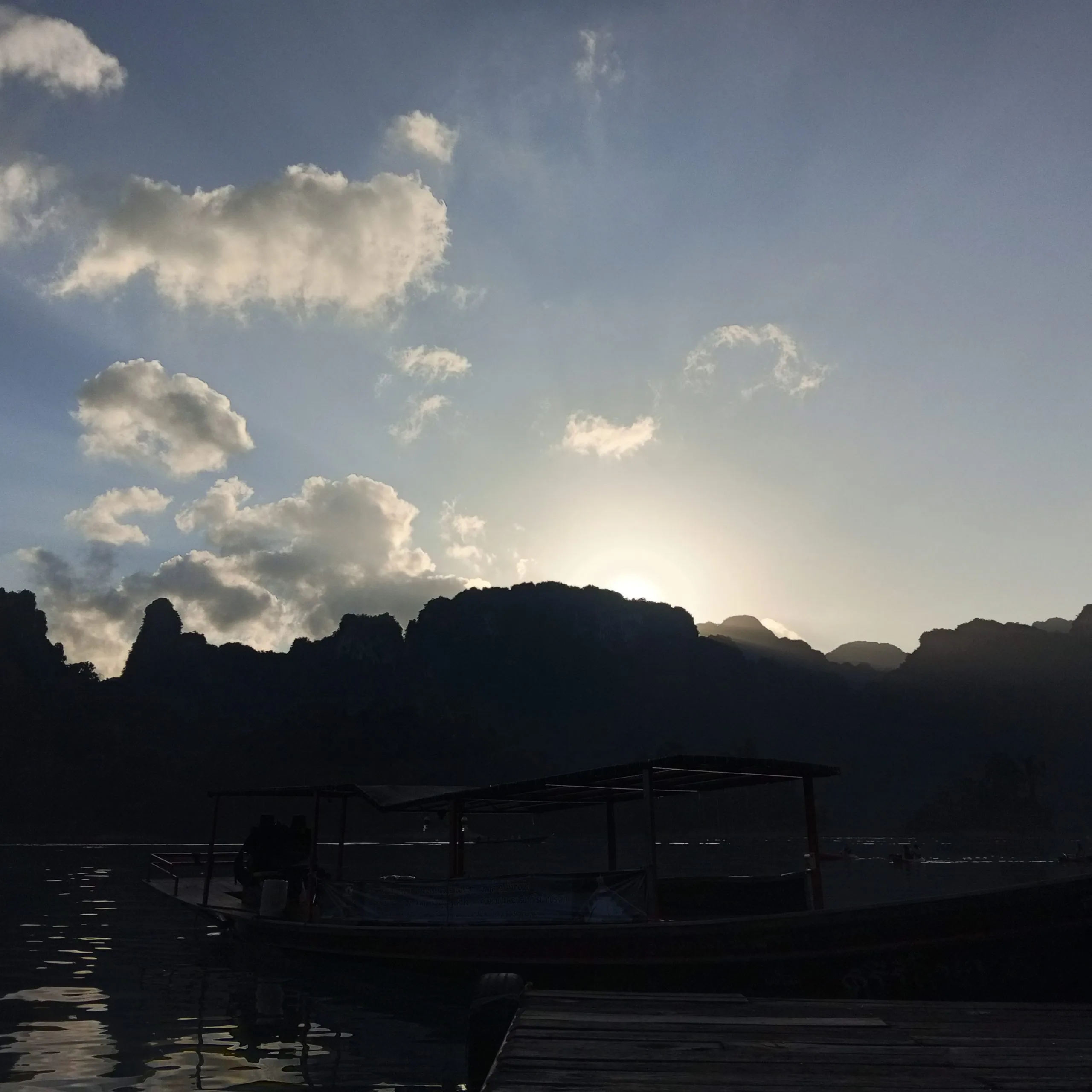 A longtail boat silhouetted against the sunrise beside limestone mountains on Cheow Lan Lake