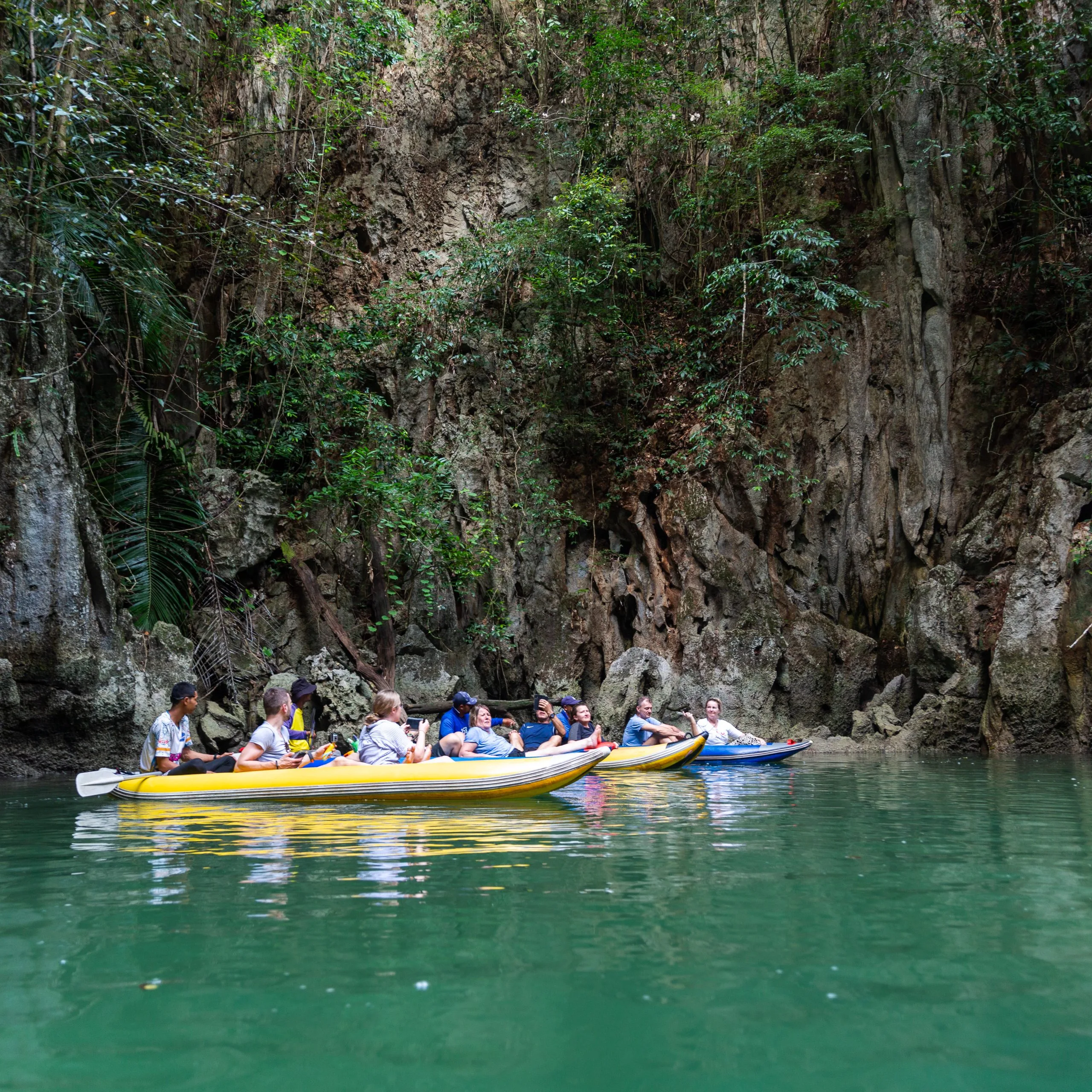 Guests paddle a yellow canoe across an emerald lagoon below jungle-covered limestone cliffs.