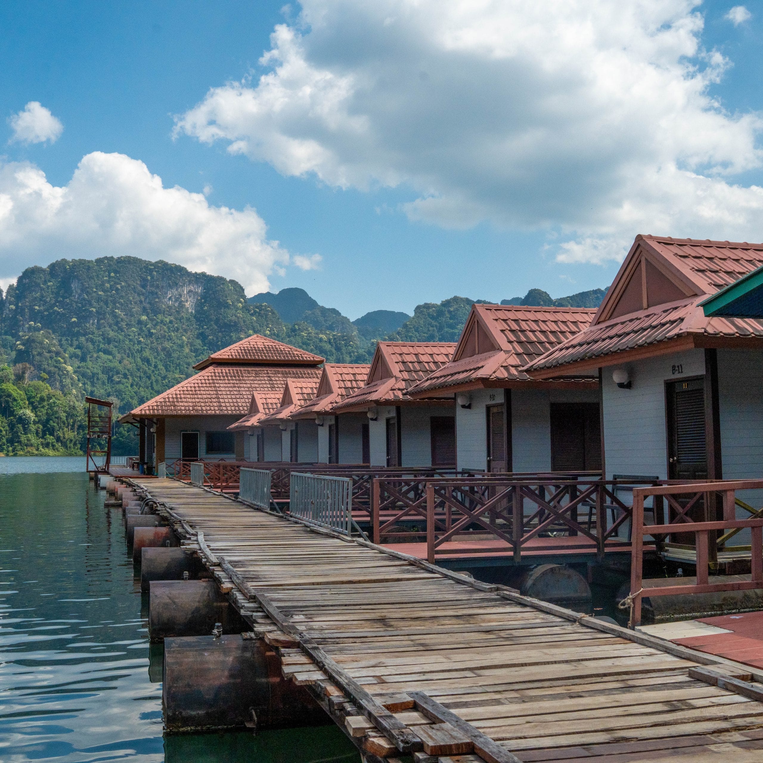 A wooden walkway running past red-roofed floating bungalows towards limestone mountains on Cheow Lan Lake