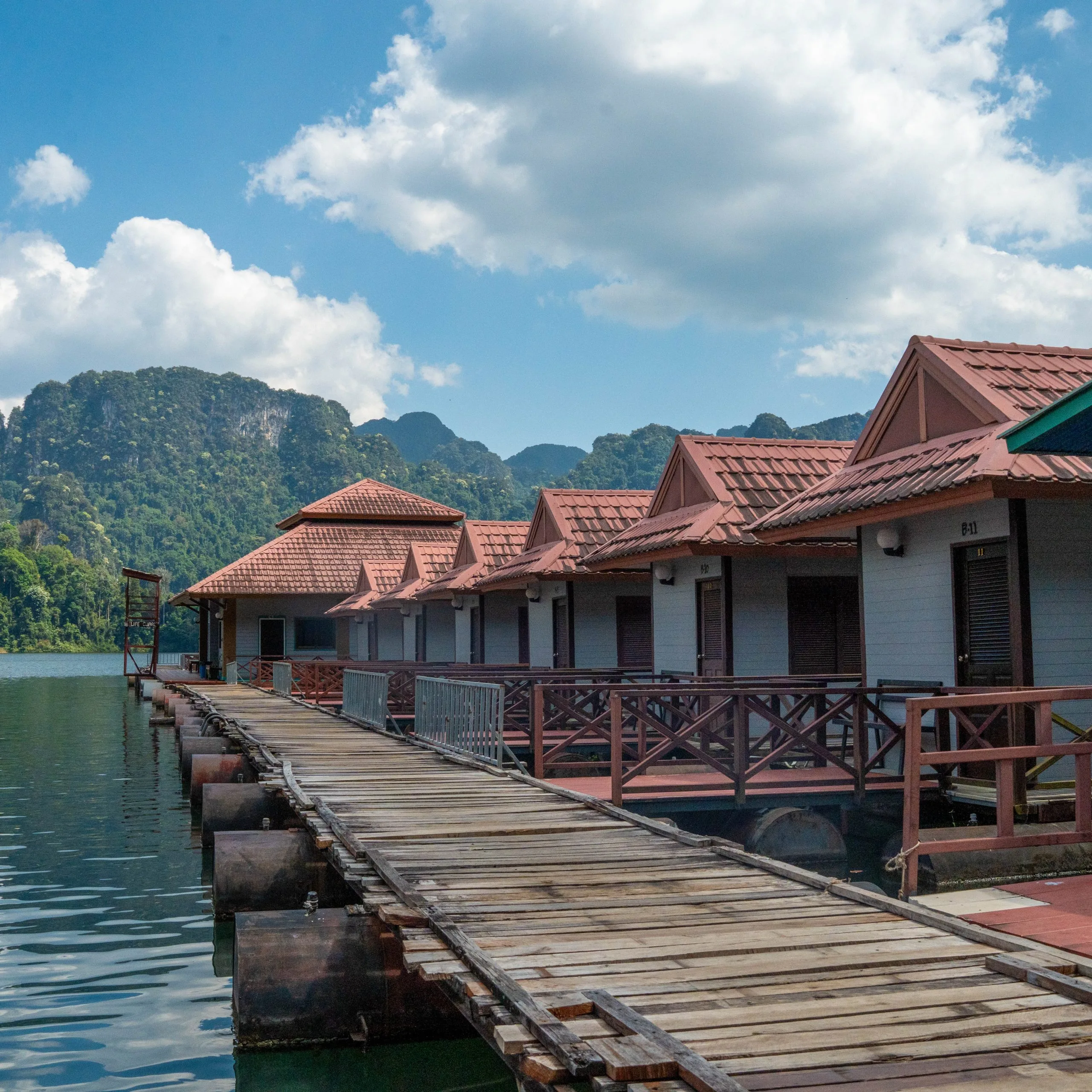 A wooden walkway running past red-roofed floating bungalows towards limestone mountains on Cheow Lan Lake