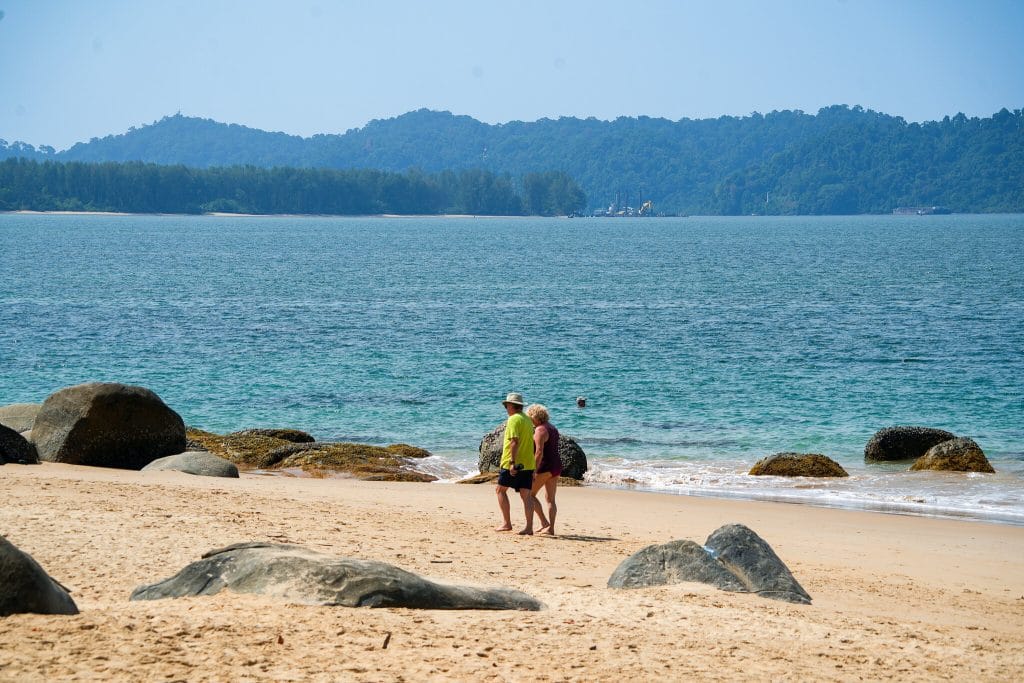 Two people walking along a quiet Khao Lak beach between granite boulders, with a forested headland behind