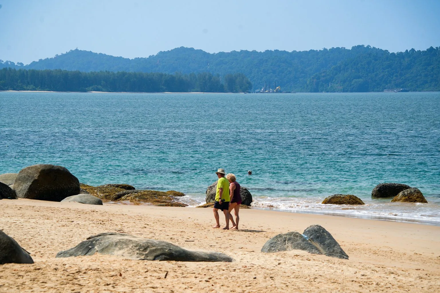 Two people walking along a quiet Khao Lak beach between granite boulders, with a forested headland behind