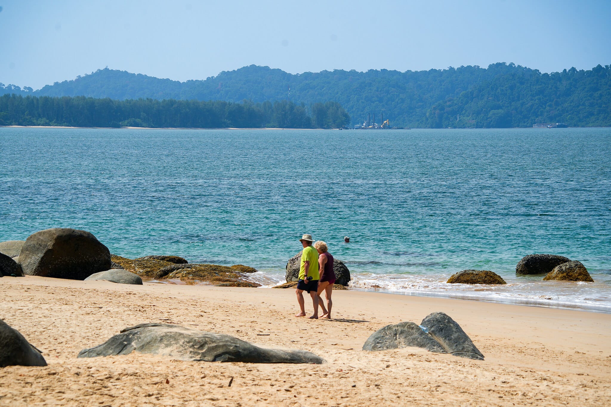 Deux personnes marchant sur une plage tranquille avec des rochers de granit et un promontoire boisé près de Khao Lak
