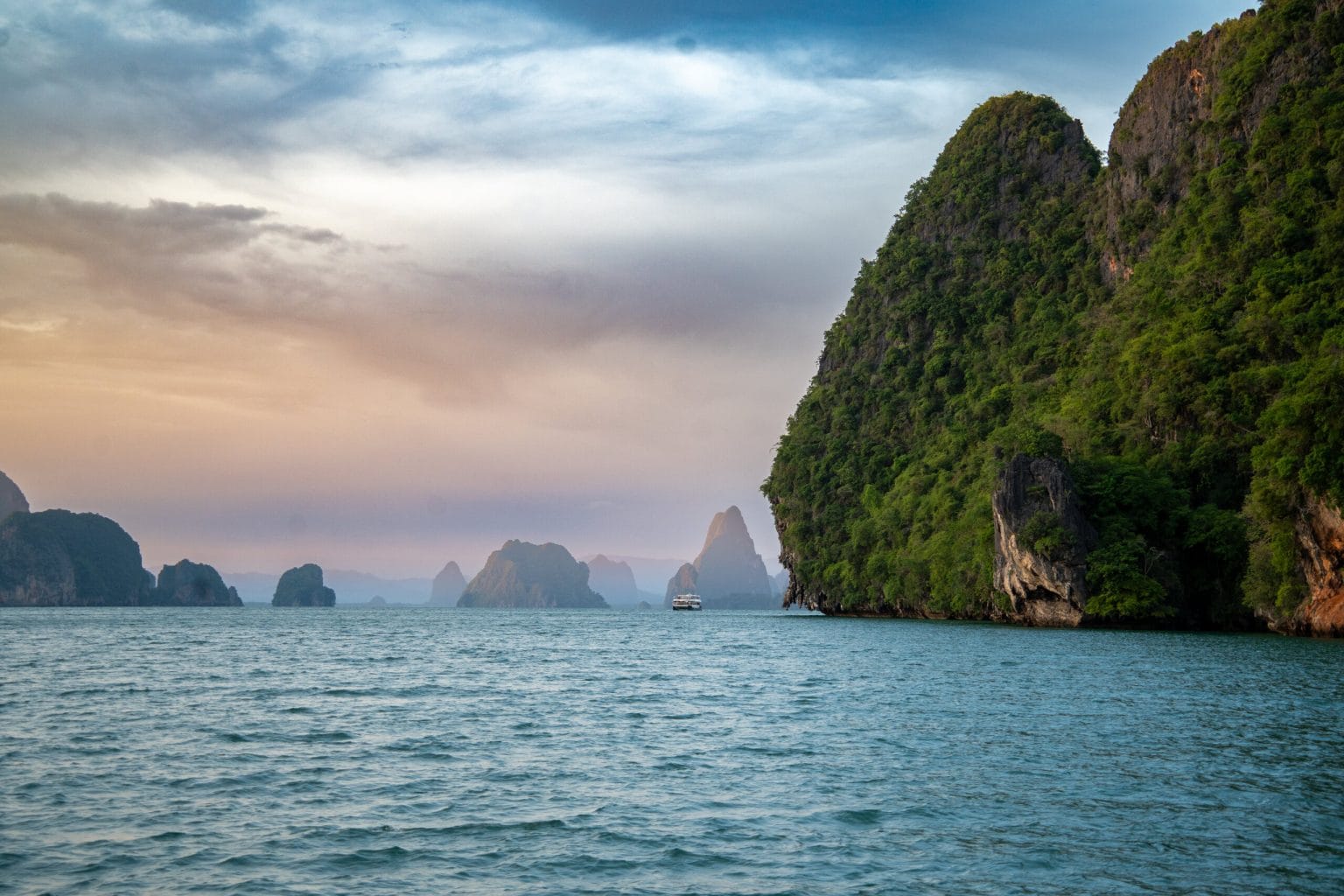 Limestone towers of Phang Nga Bay fading into the haze at dusk, with a single boat crossing the bay below a sheer, jungle-covered cliff