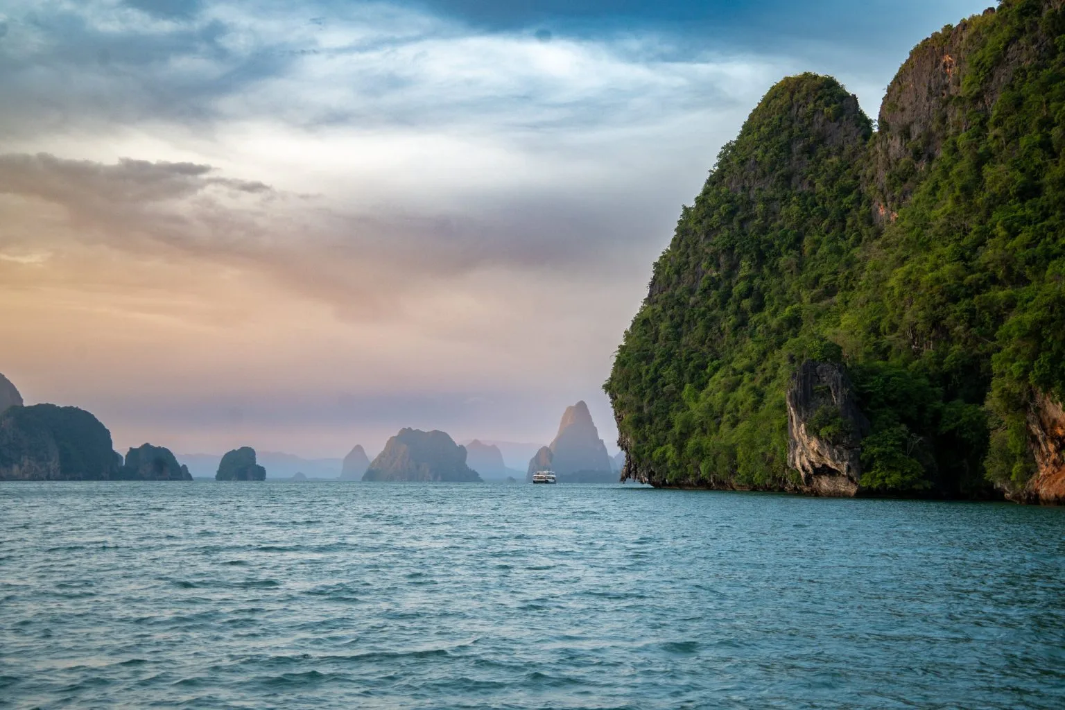 Limestone towers of Phang Nga Bay fading into the haze at dusk, with a single boat crossing the bay below a sheer, jungle-covered cliff